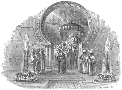 A man walks, head bowed, through an arch, with three men flanked on either side of it, while other men are in a group behind him