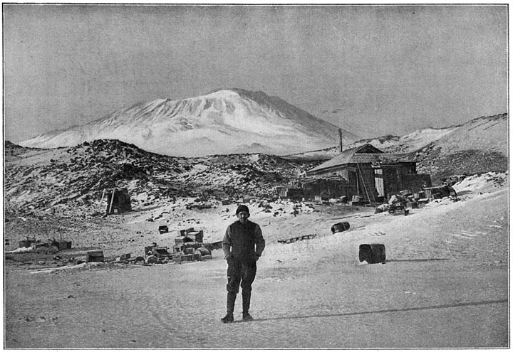 Het winterkwartier bij kaap Royds. Op den achtergrond de groote vulkaan Erebus. Luitenant Shackleton staat op den voorgrond. De lengte van zijn schaduw wijst aan, hoe laag de zon boven den horizon staat.