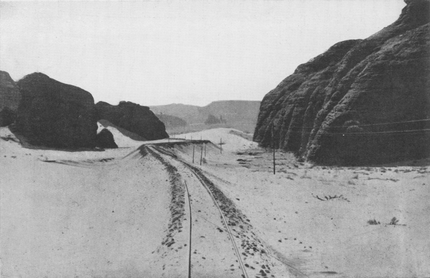 Photo wide shot of rail tracks in sand receding into the distance; stone hills border on each side