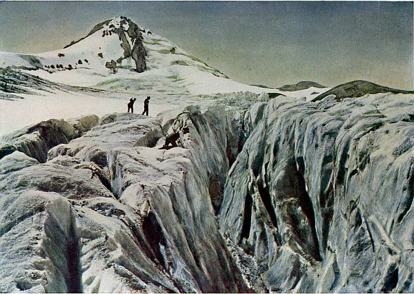 Three men, one on hands and knees looking into crevasse